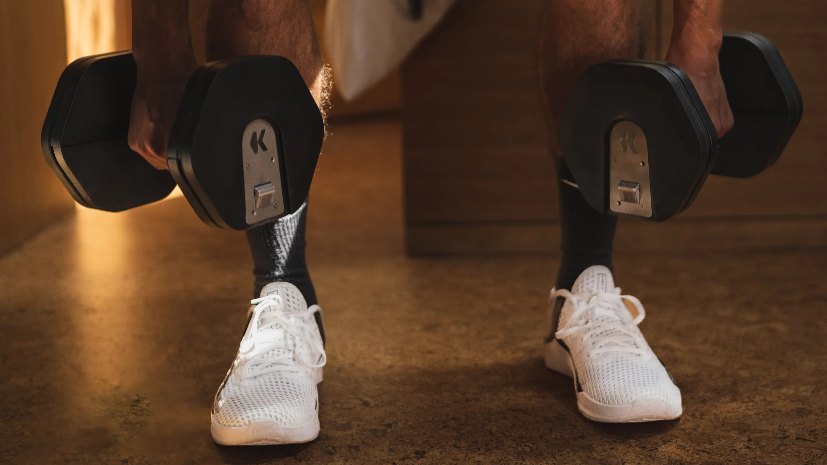 A man carrying a pair of Kabata dumbbells across a floor at home, the K mark on each end plate
