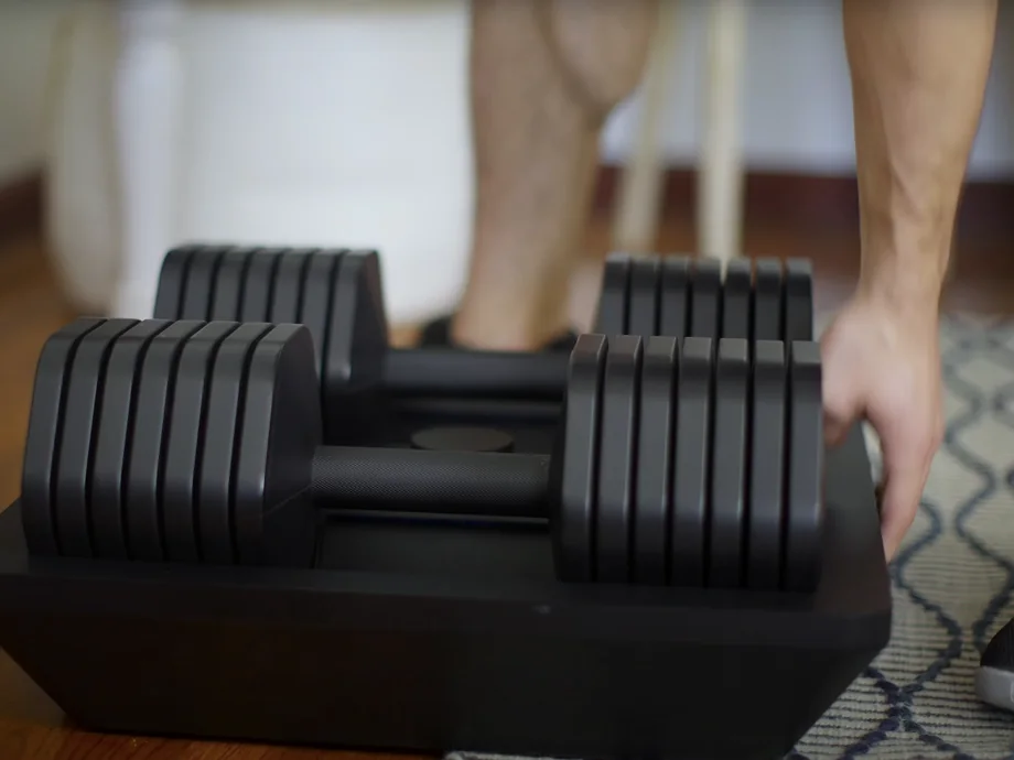 A hand lifting a Kabata dumbbell off its base on a rug at home