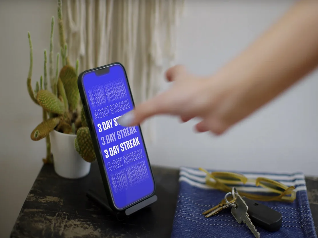 A finger tapping a phone on a desk stand, the screen filled with repeating 3 day streak lines on blue