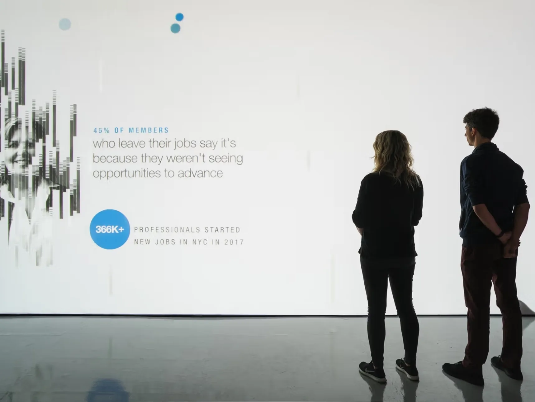 Two people reading a Talent Intelligence statistic projected the height of a gallery wall, the strand portrait resolving at its left edge.
